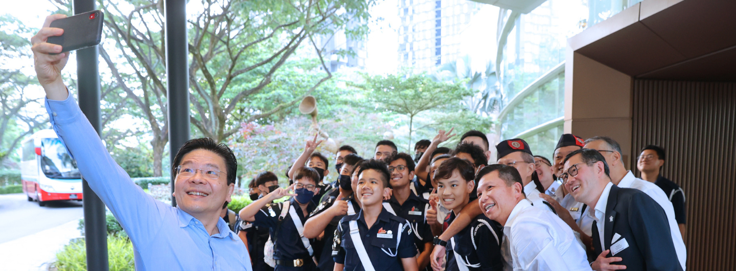 Man takes a selfie with a group outside by trees. Some wear uniform, some wear business attire.